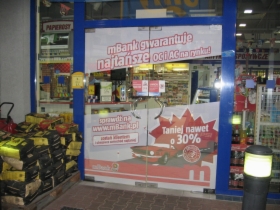 Wejście do placówki z reklamą mBanku gwarantującą najtańsze OC/AC na rynku, oklejone folią z czerwonym samochodem i napisem 'Taniej nawet o 30%', obok stos drewna opałowego i żółta skrzynka pocztowa.